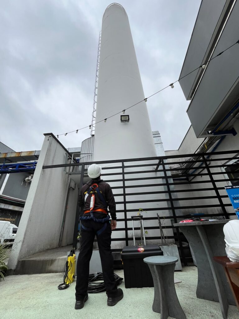 Técnico da AJM com capacete e cinto de segurança diante de estrutura elevada