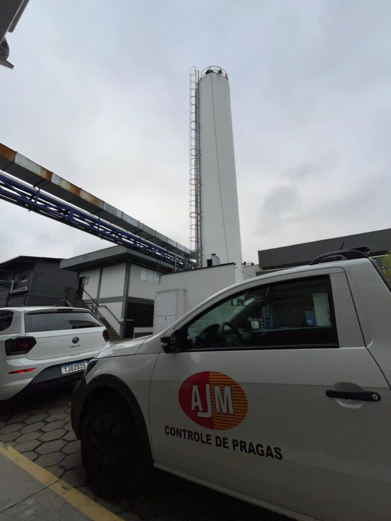 Veículo da AJM Controle de Pragas em área industrial com estrutura elevada ao fundo